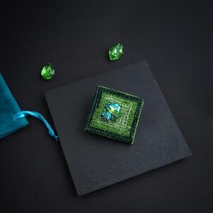 May include: A square, green beaded brooch with a central cluster of iridescent crystals. The brooch is set on a black square, with two loose green crystals and a teal pouch in the background. The brooch measures approximately 1.5 inches square.