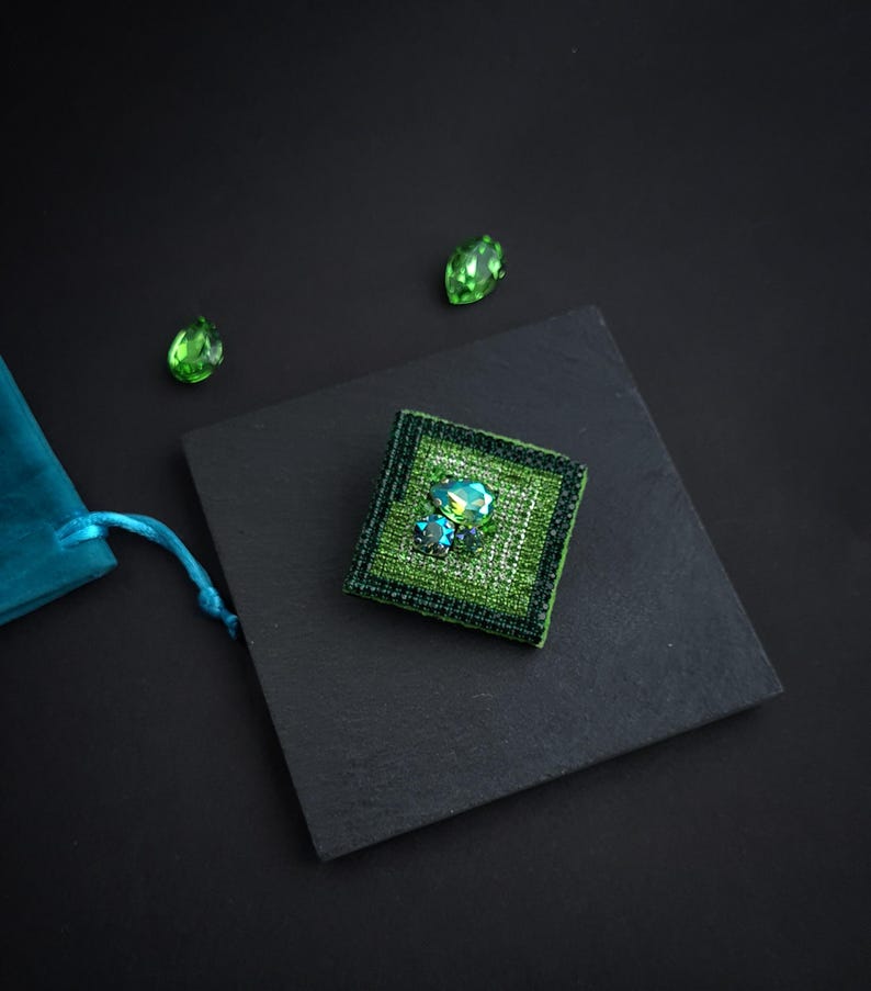 May include: A square, green beaded brooch with a central cluster of iridescent crystals. The brooch is set on a black square, with two loose green crystals and a teal pouch in the background. The brooch measures approximately 1.5 inches square.