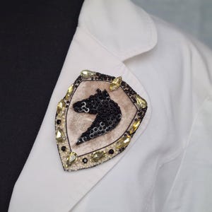 Op de afbeelding: Decoratieve broche met een zwart paardenhoofd ontwerp in een schildvorm. De broche is versierd met zwarte pailletten, goudkleurige kristallen en een beige fluwelen achtergrond. Het is vastgemaakt aan een witte blazer.