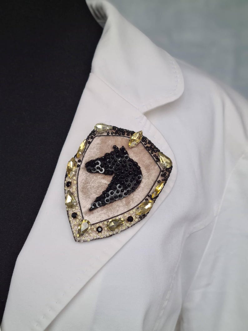 Op de afbeelding: Decoratieve broche met een zwart paardenhoofd ontwerp in een schildvorm. De broche is versierd met zwarte pailletten, goudkleurige kristallen en een beige fluwelen achtergrond. Het is vastgemaakt aan een witte blazer.