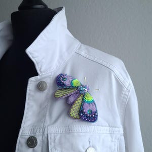 Può includere: Una spilla a forma di farfalla in perle viola e verdi con paillettes iridescenti. La spilla è appuntata su una giacca di jeans bianca.