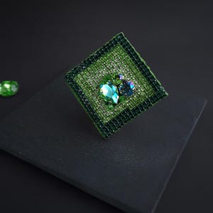 May include: A square, green brooch with a textured, woven appearance. The brooch features a large, teardrop-shaped, emerald-green gemstone and a smaller, round, blue-green gemstone, both surrounded by smaller, sparkling crystals. The brooch is set against a dark background.