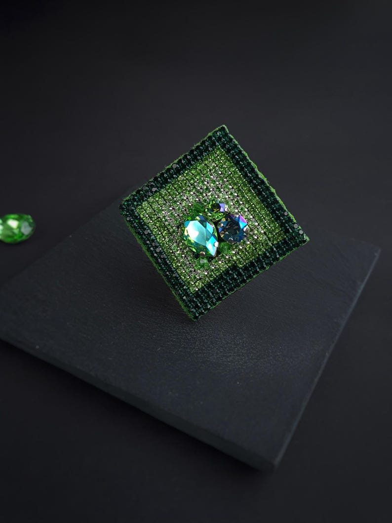 May include: A square, green brooch with a textured, woven appearance. The brooch features a large, teardrop-shaped, emerald-green gemstone and a smaller, round, blue-green gemstone, both surrounded by smaller, sparkling crystals. The brooch is set against a dark background.