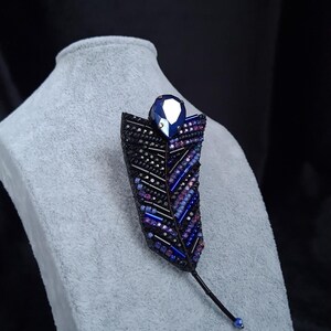 May include: A black and blue beaded brooch in the shape of a feather with a large blue crystal at the top.