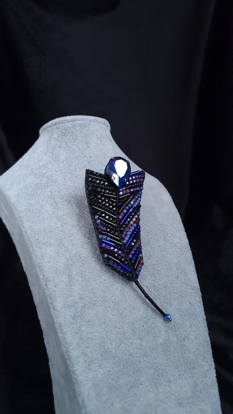 May include: A black and blue beaded brooch in the shape of a feather with a large blue crystal at the top.