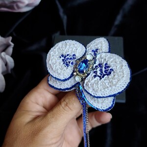 May include: A handcrafted orchid brooch featuring white and blue sequins, a central oval blue crystal, and smaller clear rhinestones. The petals are edged in deep blue thread, and a long, thin, blue beaded fringe hangs from the base of the flower.