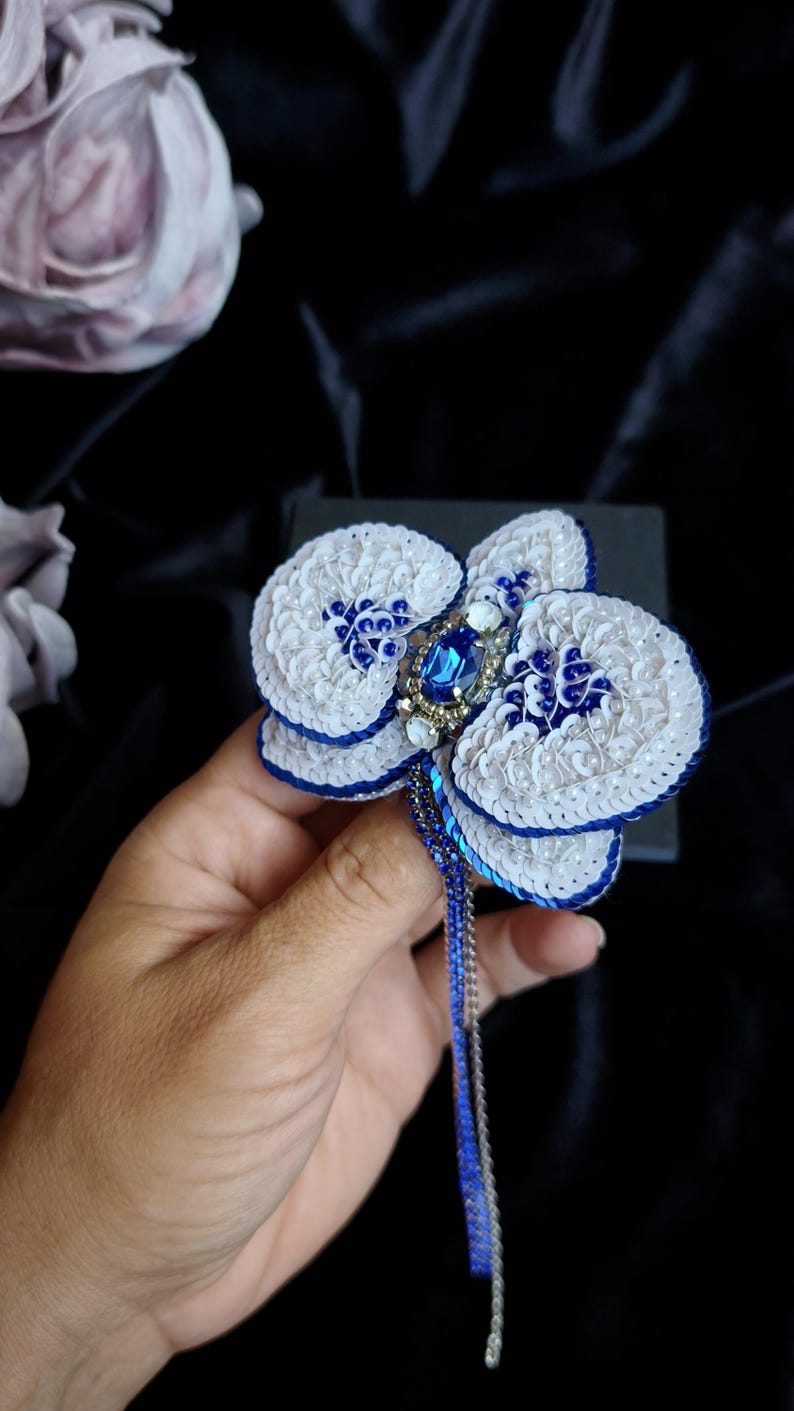May include: A handcrafted orchid brooch featuring white and blue sequins, a central oval blue crystal, and smaller clear rhinestones. The petals are edged in deep blue thread, and a long, thin, blue beaded fringe hangs from the base of the flower.