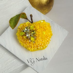 May include: A handmade yellow sequin apple brooch with a green leaf and stem, embellished with crystals. The brooch is on a white square, with the designer's name, "V.G. Natalia," visible.