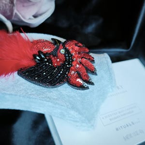 May include: A handcrafted brooch shaped like a bird, featuring vibrant red and black beads, sequins, and soft red feathers. The piece is displayed on a light gray fabric stand next to a white box with the text RITUALS and A SCENTED WAX CANDLE.