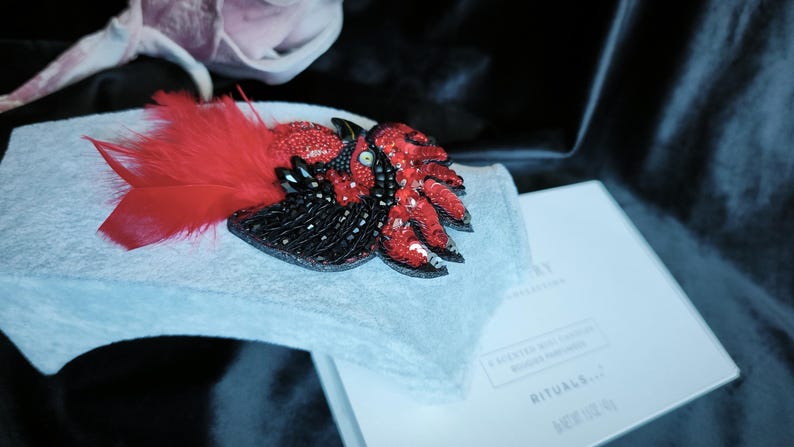 May include: A handcrafted brooch shaped like a bird, featuring vibrant red and black beads, sequins, and soft red feathers. The piece is displayed on a light gray fabric stand next to a white box with the text RITUALS and A SCENTED WAX CANDLE.