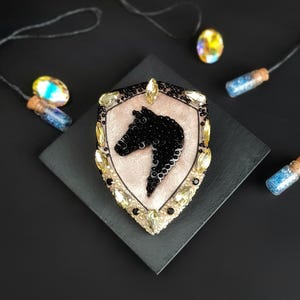 Op de afbeelding: Een decoratieve broche in de vorm van een schild, met een zwart paardenhoofd ontwerp. De broche is versierd met zwarte pailletten en gele edelstenen, tegen een lichtroze achtergrond. De broche ligt op een zwarte vierkant, met decoratieve flesjes op de achtergrond.