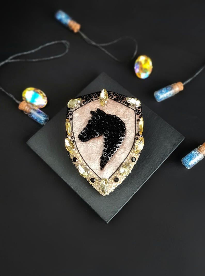 Op de afbeelding: Een decoratieve broche in de vorm van een schild, met een zwart paardenhoofd ontwerp. De broche is versierd met zwarte pailletten en gele edelstenen, tegen een lichtroze achtergrond. De broche ligt op een zwarte vierkant, met decoratieve flesjes op de achtergrond.