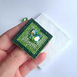May include: A square, green brooch with a sparkling, jewel-like design. The brooch features a central cluster of iridescent stones and is surrounded by a border of smaller, glittering accents. The piece is held in a hand against a white background.