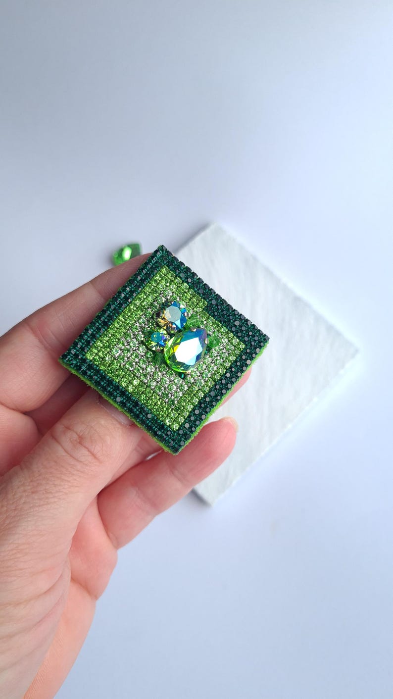 May include: A square, green brooch with a sparkling, jewel-like design. The brooch features a central cluster of iridescent stones and is surrounded by a border of smaller, glittering accents. The piece is held in a hand against a white background.