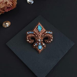 Op de afbeelding: Fleur-de-lis broche met een bronskleurige metalen basis, versierd met blauwe en heldere kristallen. De broche ligt op een donkere vierkante ondergrond, met gedroogde bloemen en losse kristallen op de achtergrond. Een decoratief accessoire.