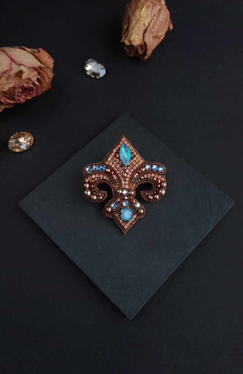 Op de afbeelding: Fleur-de-lis broche met een bronskleurige metalen basis, versierd met blauwe en heldere kristallen. De broche ligt op een donkere vierkante ondergrond, met gedroogde bloemen en losse kristallen op de achtergrond. Een decoratief accessoire.