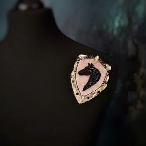 Op de afbeelding: Decoratieve broche met een schildvorm en een silhouet van een paardenhoofd. De broche heeft een roze achtergrond, een zwart paardenhoofd en is versierd met zwarte pailletten en heldere kristallen. Bevestigd aan een zwarte mannequin.