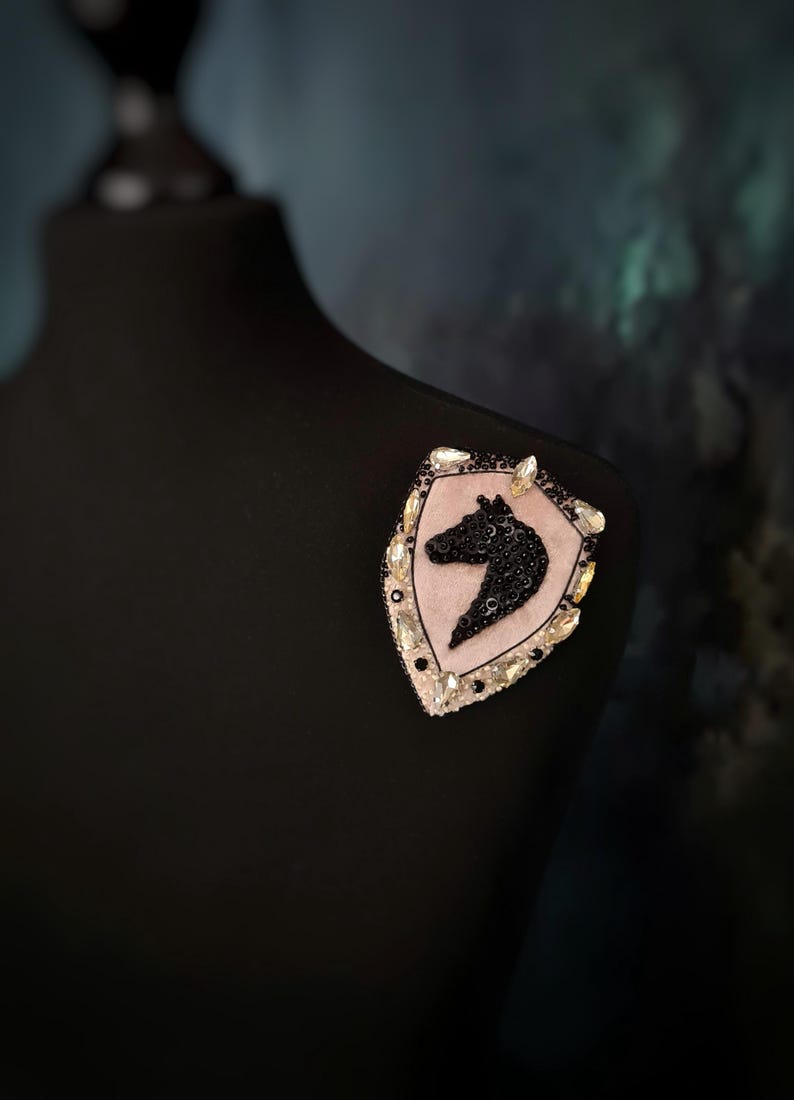 Op de afbeelding: Decoratieve broche met een schildvorm en een silhouet van een paardenhoofd. De broche heeft een roze achtergrond, een zwart paardenhoofd en is versierd met zwarte pailletten en heldere kristallen. Bevestigd aan een zwarte mannequin.