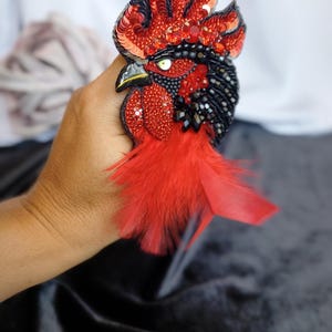 May include: A handcrafted rooster brooch featuring vibrant red and black sequins, beads, and crystals. The design includes a detailed eye, a pointed beak, and soft red feathers extending from the base, creating a textured, layered appearance.