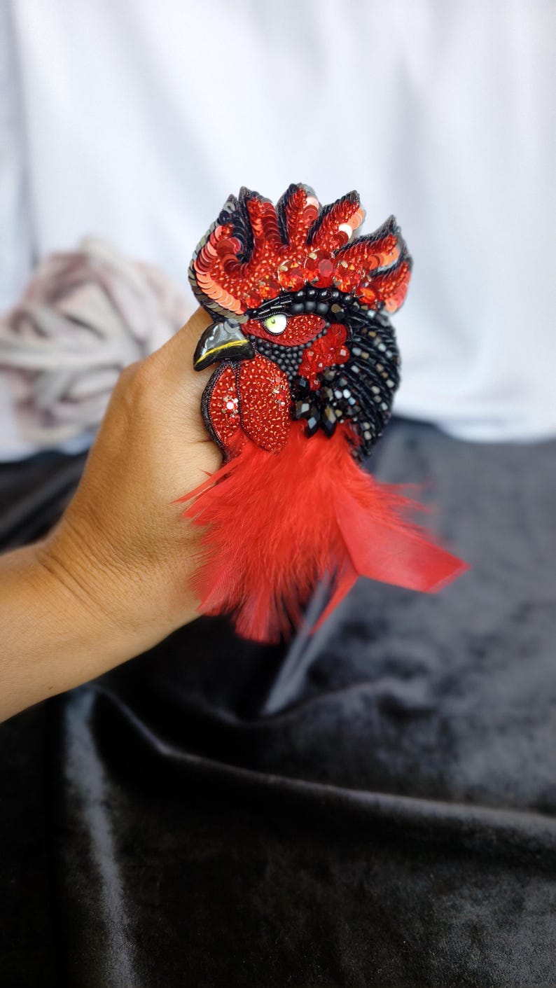 May include: A handcrafted rooster brooch featuring vibrant red and black sequins, beads, and crystals. The design includes a detailed eye, a pointed beak, and soft red feathers extending from the base, creating a textured, layered appearance.