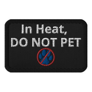 以下が含まれることがあります： 黒い刺繍パッチに、白い文字で「In Heat, Do Not Pet」と書いてあり、赤い円の中に青い肉球の跡があり、その上に斜めの赤い線が引かれている。