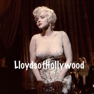 May include: A woman with blonde hair and red lipstick wearing a sequined dress and jewelry. The image has the text "LloydsofHollywood" in white. The background includes a dark curtain and musical instruments.