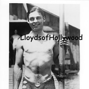 Puede incluir: Fotografía vintage en blanco y negro de un hombre sonriente en traje de baño, sosteniendo un poste. Los pantalones cortos tienen detalles recortados. La imagen está marcada con el texto "LloydsofHollywood".