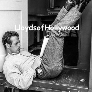 Puede incluir: Una fotografía en blanco y negro de un hombre acostado de espaldas con las piernas en el aire. Lleva pantalones vaqueros y botas. Tiene un papel en la mano y un cigarrillo en la boca.