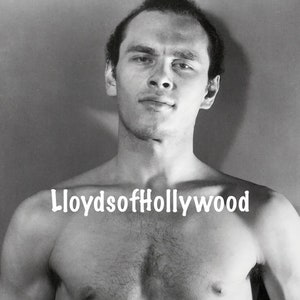 May include: Black and white photo of a shirtless man with short hair. The man is looking directly at the camera. The photo is taken from the chest up.