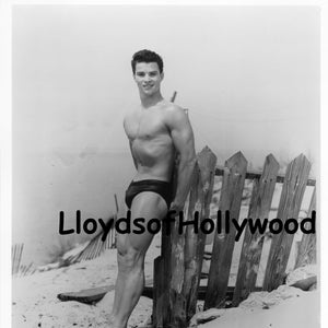 Puede incluir: Una fotografía en blanco y negro de un hombre de pie en una playa, apoyado en una valla de madera. Lleva un bañador y tiene un físico musculoso.