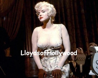 May include: A woman with blonde hair wearing a sparkly, gold and white dress. She is sitting on a chair with a microphone in front of her. The background is a dark stage with curtains.