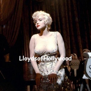 May include: A woman with blonde hair wearing a sparkly, gold and white dress. She is sitting on a chair with a microphone in front of her. The background is a dark stage with curtains.
