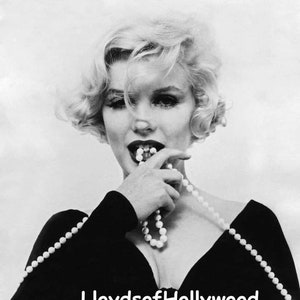 May include: A black and white photo of a woman wearing a black dress and a pearl necklace. She is biting on the necklace and looking at the camera. The text "Lloyds of Hollywood" is visible at the bottom of the image.
