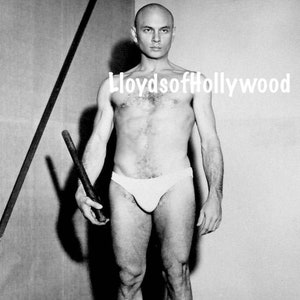 May include: Black and white photograph of a bald person wearing white briefs and holding a dark wooden rod. The person is standing in a corner with a light-colored wall in the background. The watermark "LloydsofHollywood" is visible.