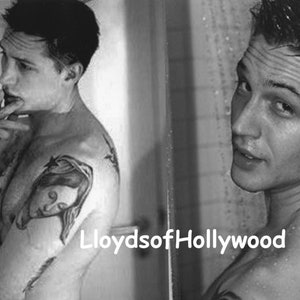 May include: Black and white image of a person with tattoos on their shoulder and arm. The person is looking towards the camera. The text "LloydsofHollywood" is visible in the image.