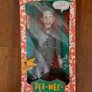 Puede incluir: Un muñeco parlante vintage de Pee-wee Herman con un traje gris y una corbata de lazo roja. El muñeco está empaquetado en una caja con las palabras "Talking Pee-wee Herman" y "Pee-wee's Playhouse" en ella. La caja es naranja y verde con un diseño de dibujos animados.