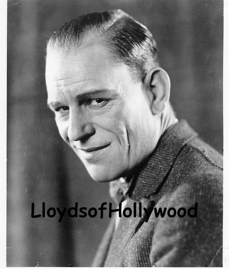 May include: Black and white portrait photograph of a man with a receding hairline, wearing a patterned suit jacket. The man is smiling and looking to the left of the camera. The text "LloydsofHollywood" is visible in the bottom left corner of the image.