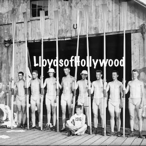 Puede incluir: Una fotografía en blanco y negro de un equipo de remo posando frente a un cobertizo para botes. El equipo sostiene remos y lleva trajes de baño. La fotografía está fechada en 1916.