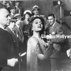 May include: A black and white photo of a group of people on a film set. A woman in a sparkly dress is speaking to the camera. The text "Lloyds of Hollywood" is visible in the image.