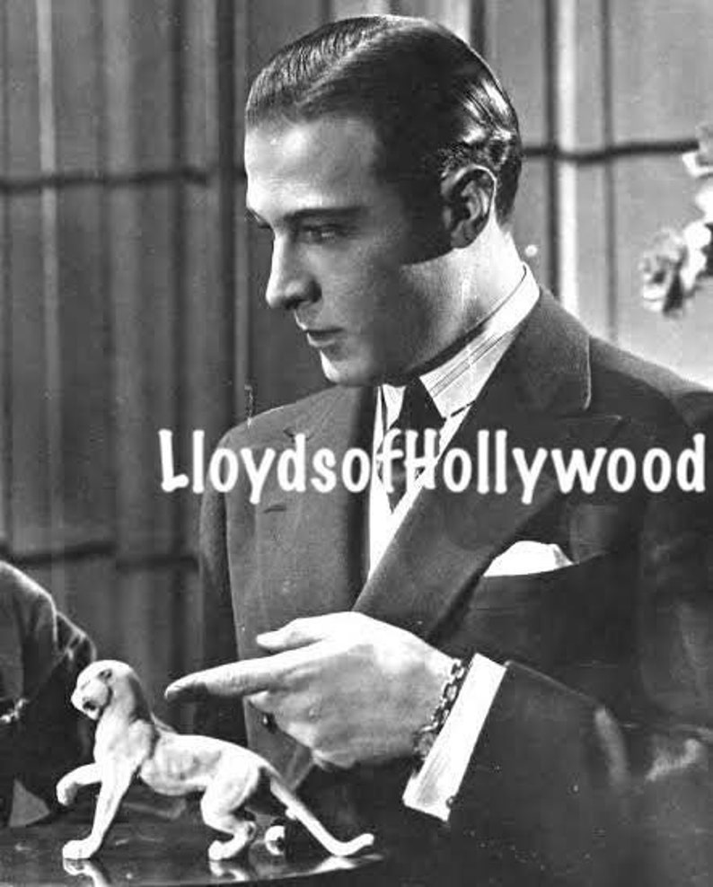 May include: A black and white photo of a man in a suit, with a serious expression on his face. He is wearing a dark suit with a white shirt and a tie. The man is sitting at a table with a small monkey figurine in front of him. The photo is captioned "Lloyds of Hollywood".