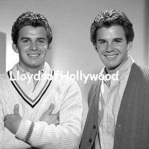 May include: A black and white photograph featuring two individuals with dark, wavy hair, both smiling. One wears a cable-knit V-neck sweater with striped trim, while the other wears a button-down shirt under a knit cardigan. Text reads Lloyds of Hollywood.