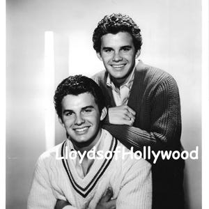 May include: A black and white photograph featuring two individuals with dark, wavy hair. One wears a V-neck sweater with striped trim, while the other wears a textured cardigan over a collared shirt. The text Lloyds of Hollywood is visible.