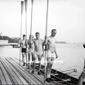 Puede incluir: Fotografía en blanco y negro de un equipo de remo de pie en un muelle de madera, sosteniendo remos. Los hombres visten pantalones cortos. Un hombre con un megáfono está cerca. El fondo muestra agua y un cobertizo para botes.