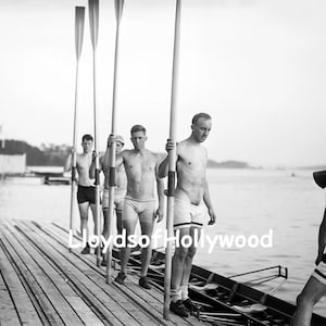 Puede incluir: Una fotografía en blanco y negro de un equipo de remo en un muelle de madera. El equipo está formado por cinco hombres, todos sin camisa y con pantalones de baño. Están sosteniendo remos y mirando a la cámara. El muelle está hecho de tablones de madera y hay agua en el fondo.