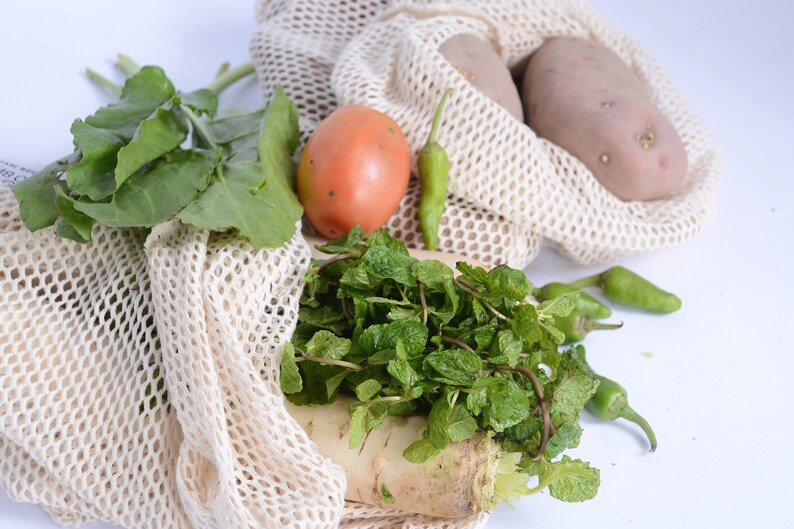 May include: A white mesh produce bag filled with fresh produce, including green leafy vegetables, a red tomato, green peppers, a bunch of mint, and a white radish. Two brown potatoes are in a separate white mesh bag.