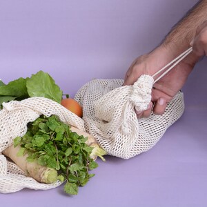 May include: A person is holding a white mesh produce bag filled with fresh vegetables, including leafy greens and a white radish. The bag is on a light purple surface.