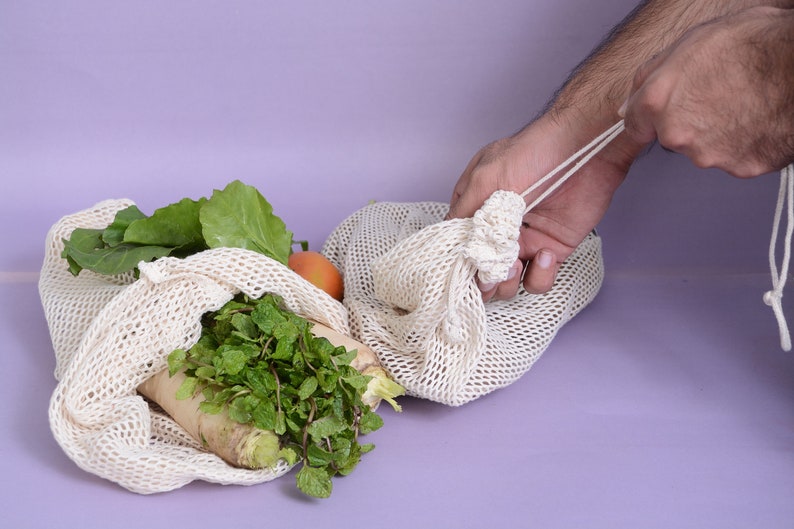 May include: A person is holding a white mesh produce bag filled with fresh vegetables, including leafy greens and a white radish. The bag is on a light purple surface.