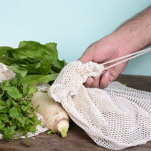 May include: A white mesh produce bag with green leafy vegetables and a white radish inside. A person's hand is holding the bag's drawstring.