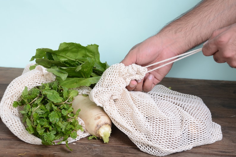 May include: A white mesh produce bag with green leafy vegetables and a white radish inside. A person's hand is holding the bag's drawstring.
