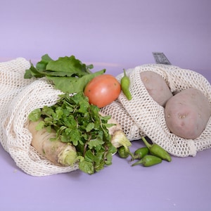 May include: Two white mesh produce bags filled with fresh produce. One bag contains a tomato, two potatoes, and green peppers. The other bag contains a bunch of green leafy herbs, a white radish, and a green leafy vegetable.